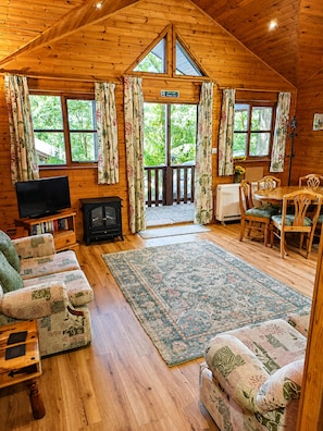 Ein Blockhaus-Interieur mit Holzbalkendecke, Kamin, Fernseher, einer Couch, einem Esstisch mit Stühlen und Blick ins Freie durch große Fenster.