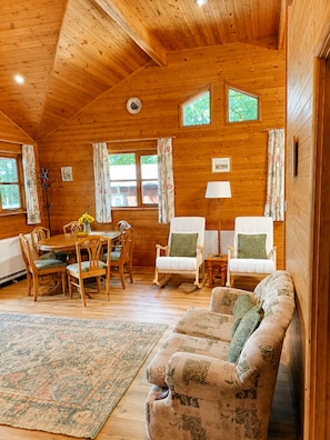 Ein gemütliches Cabin-Interieur mit Holzbalkendecke, einem Essbereich mit Tisch und Stühlen, einer Couch und einer Lampe.