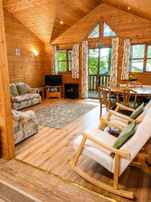 Ein Holzblock-Innenraum mit einer Couch, Sesseln, einem Fernseher, einem Kamin und großen Fenstern.