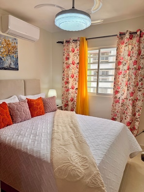A hotel room with a bed, a ceiling fan, a wall-mounted air conditioner, and a window with floral curtains. - Beachfront apartment, colonial district (Santo Domingo Este)