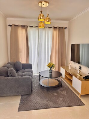 A modern living room with a grey sofa, a round black coffee table, a TV on a wooden cabinet, and a large window with curtains. - Beachfront apartment, colonial district (Santo Domingo Este)