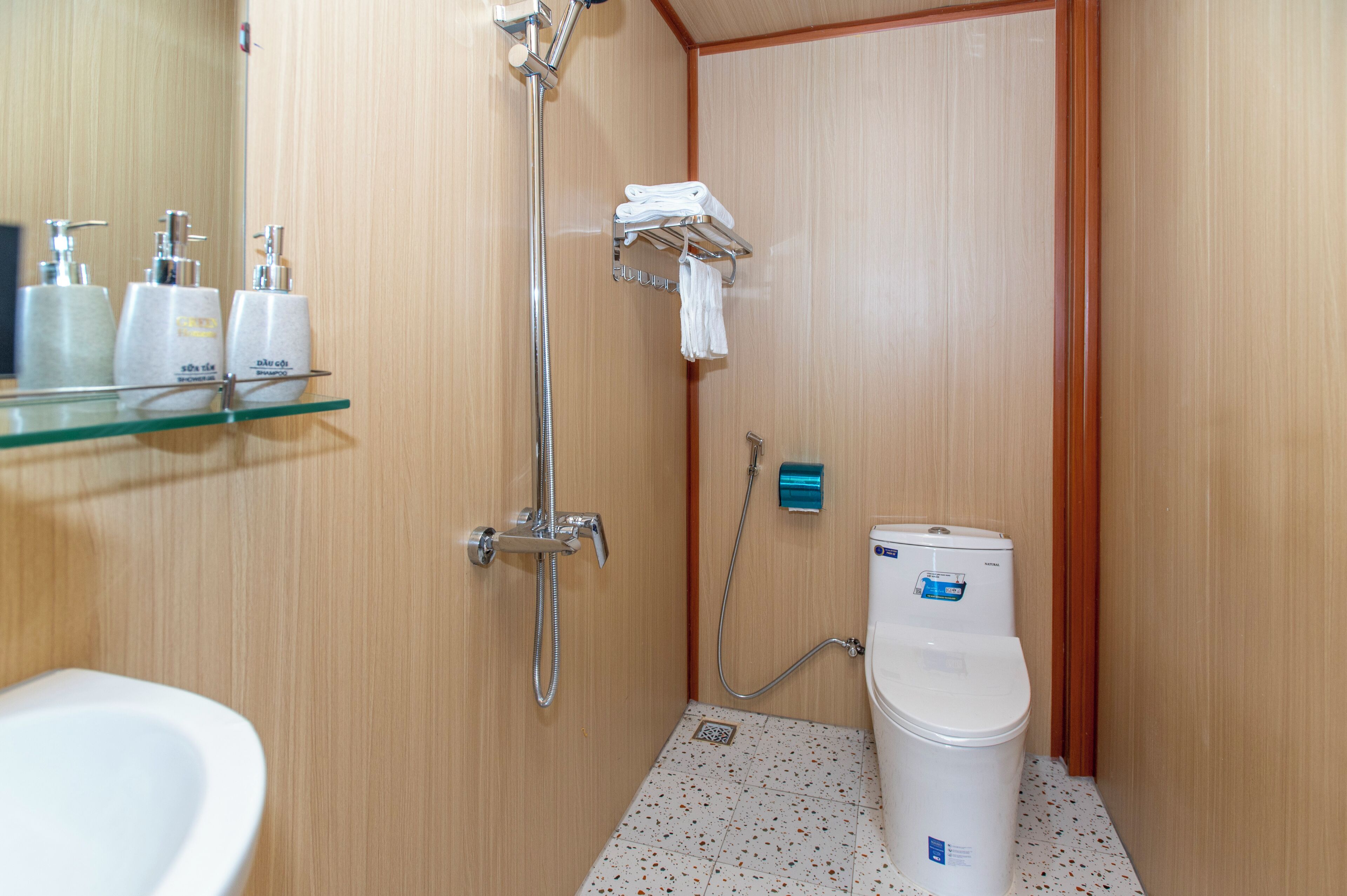 Une petite salle de bain compacte, équipée d’une douche, de toilettes et d’une étagère en verre où sont rangés les articles de toilette.