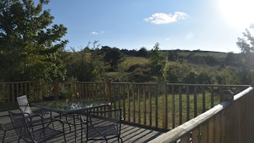 Una terraza con mesa y sillas de vidrio, con vista a un paisaje con césped, árboles y colinas.