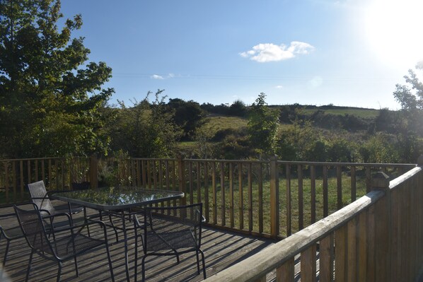 A deck with a glass table and chairs, overlooking a grassy landscape with trees and hills. - Snowdonia Riverside Cabin With Private River (Caernarfon)