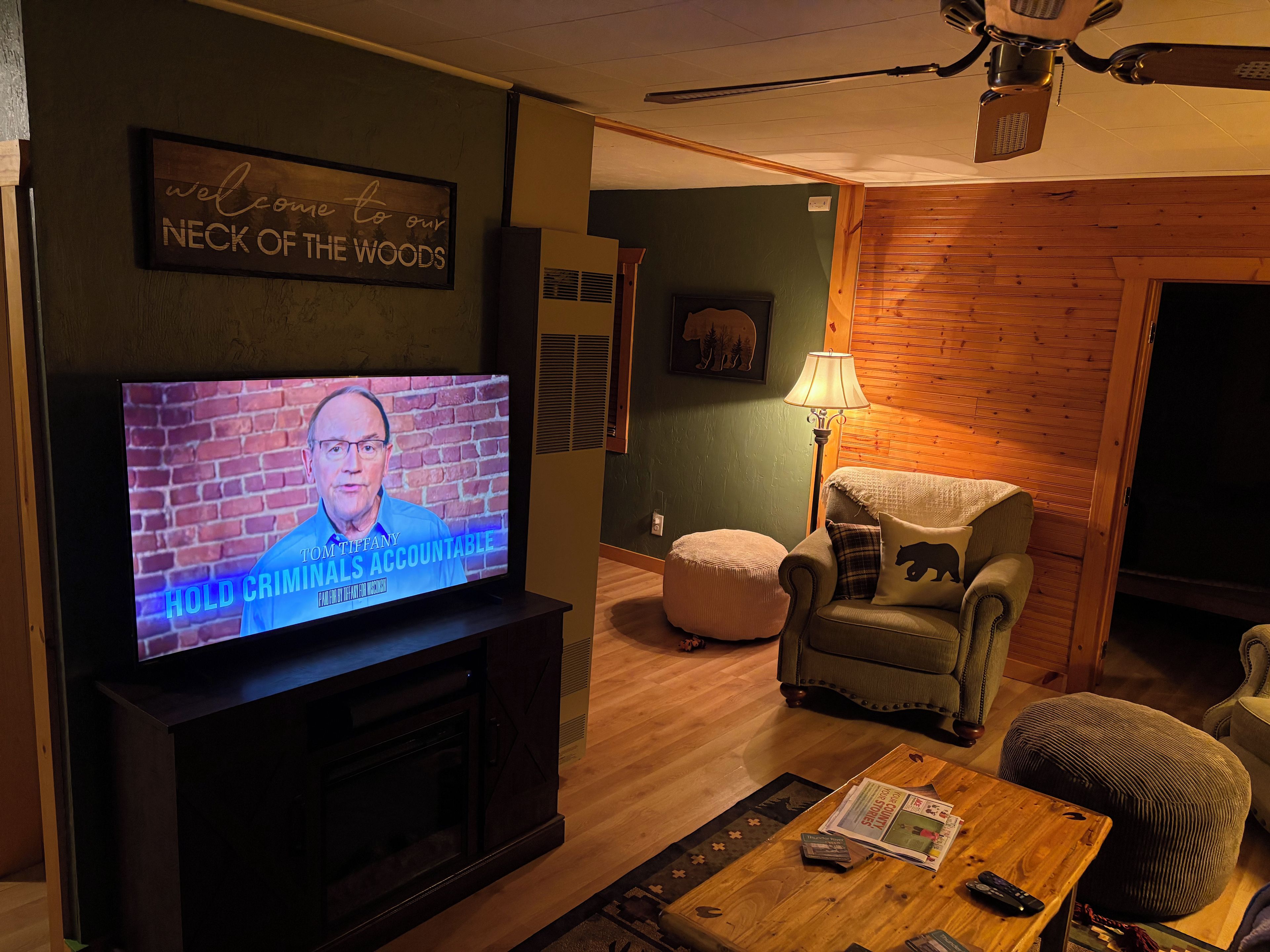 A living room with a television displaying a news program, a fireplace, a comfortable armchair, a wooden coffee table, and a wall-mounted sign.