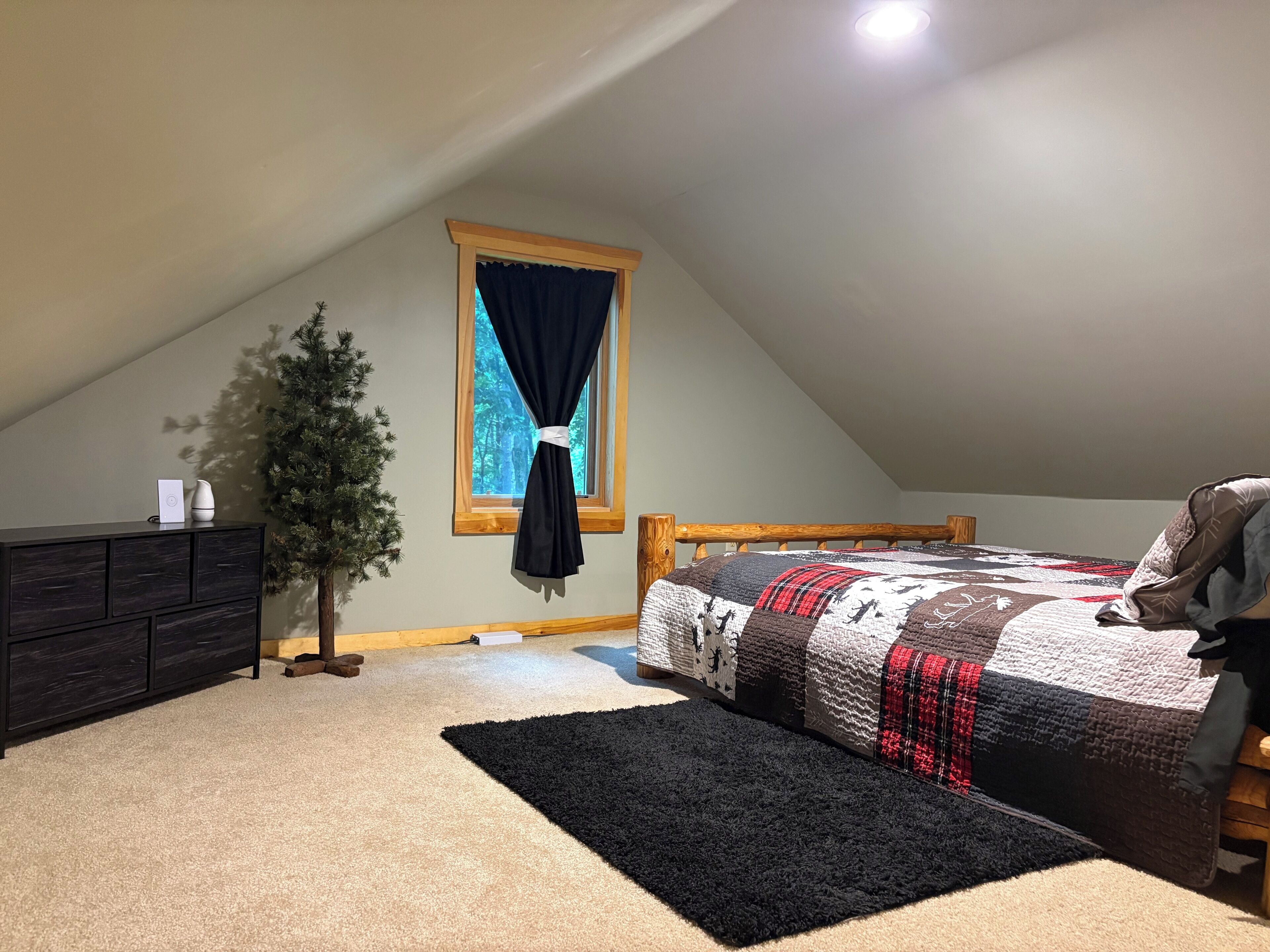 A bedroom with a sloped ceiling, a bed with a quilt, a dresser, a small Christmas tree, and a window with curtains.