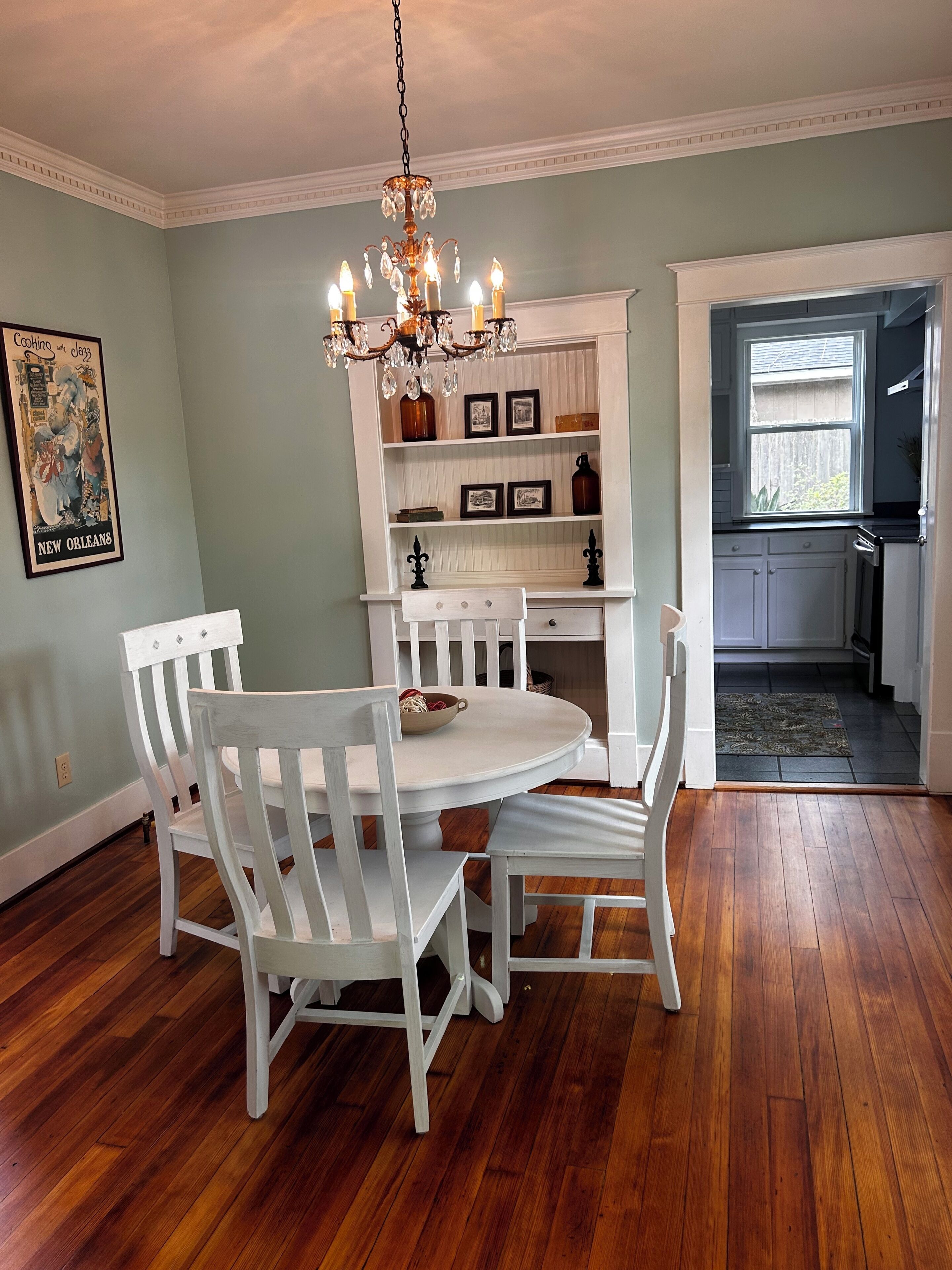 A dining area with a white round table and chairs, a chandelier, and a kitchen visible through an open door.