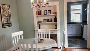 Un comedor con una mesa redonda blanca y sillas, una araña y una cocina visible a través de una puerta abierta.
