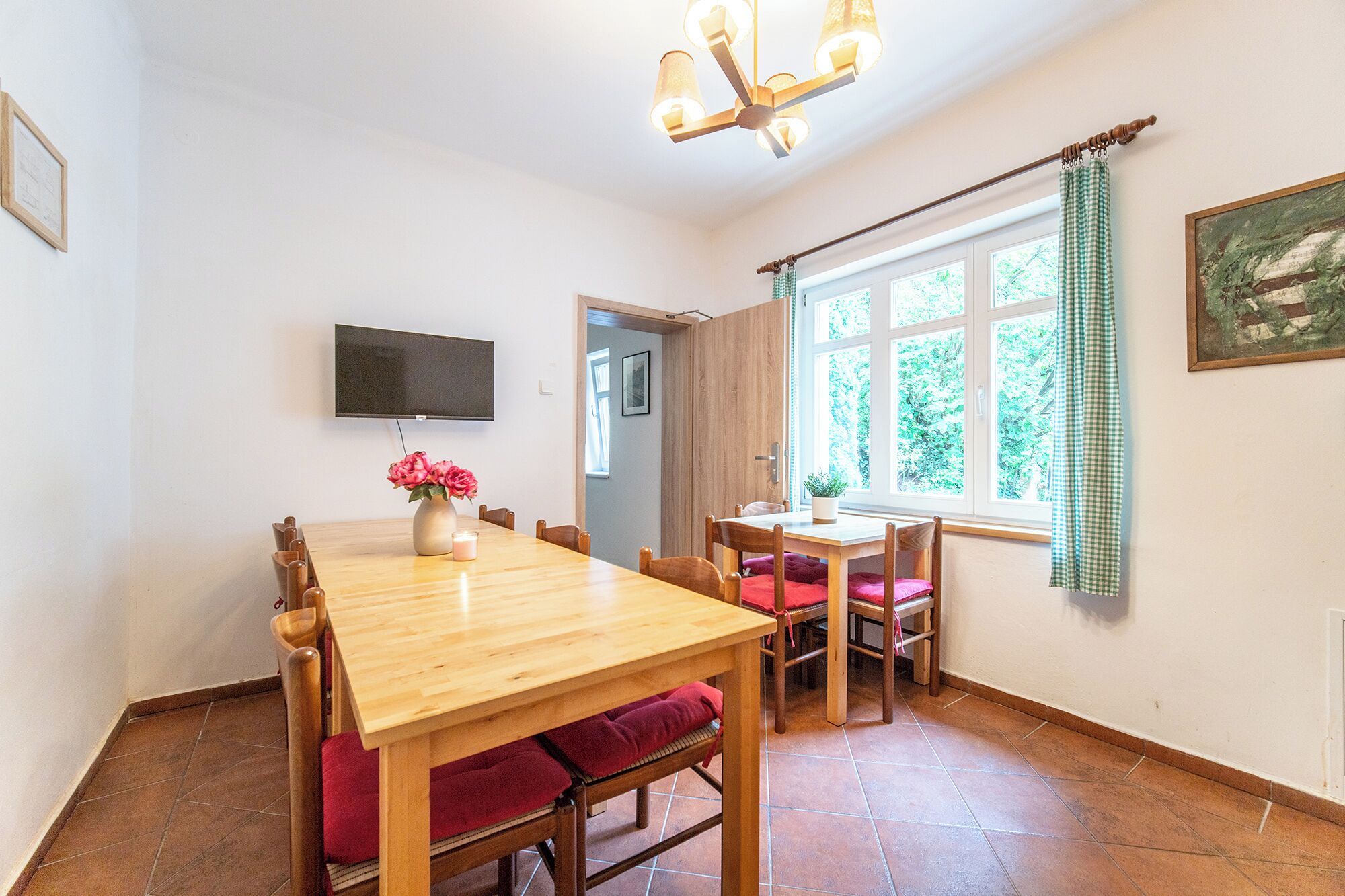 A dining area with a wooden table, chairs, a vase with flowers, a candle, and a television mounted on the wall.