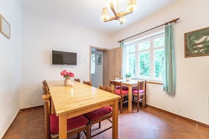 Een eethoek met een houten tafel, stoelen, een vaas met bloemen, een kaars en een aan de muur bevestigde televisie.