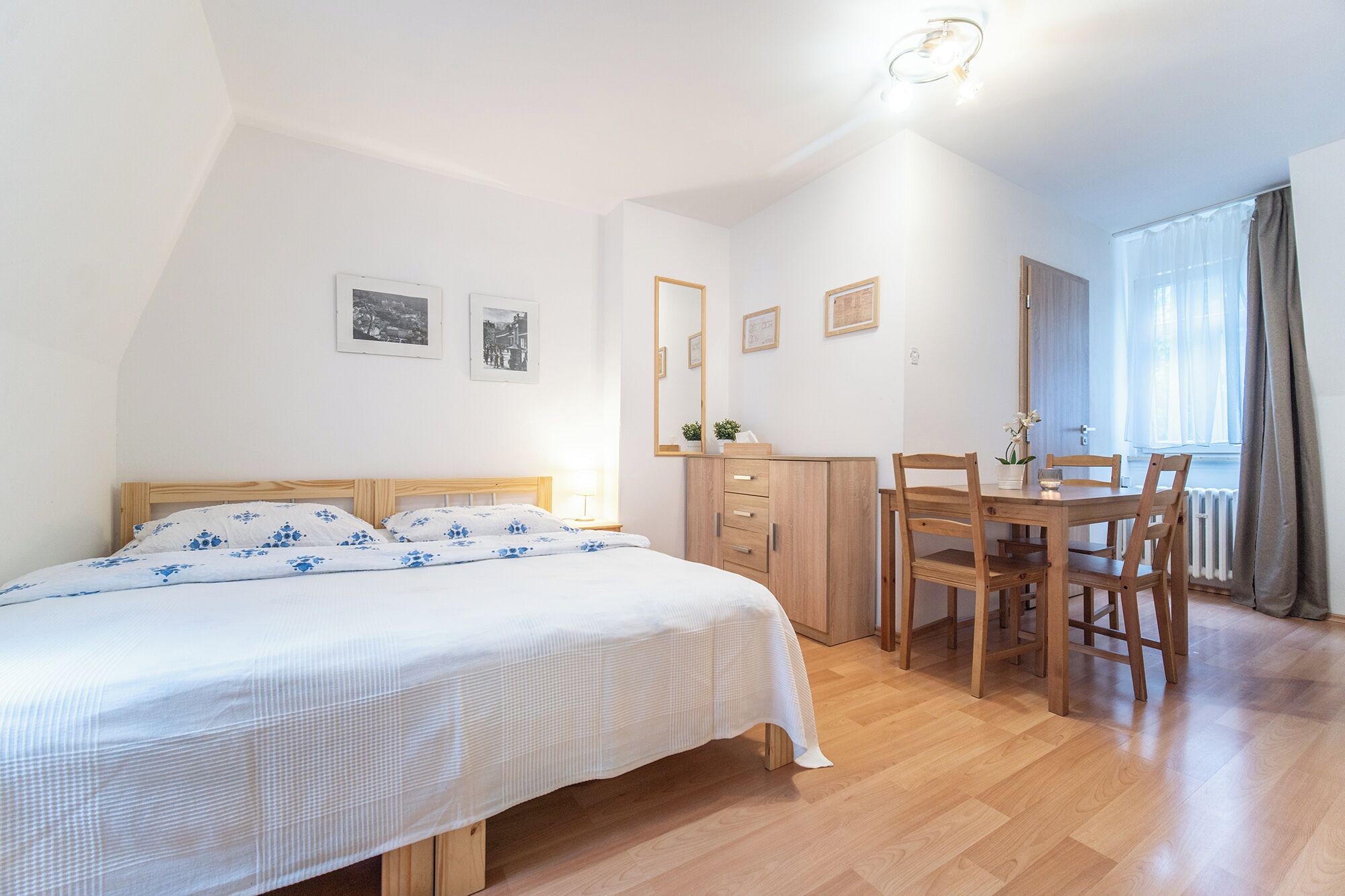 A bedroom with a bed, a wooden dresser, a dining table with chairs, and a window with curtains.