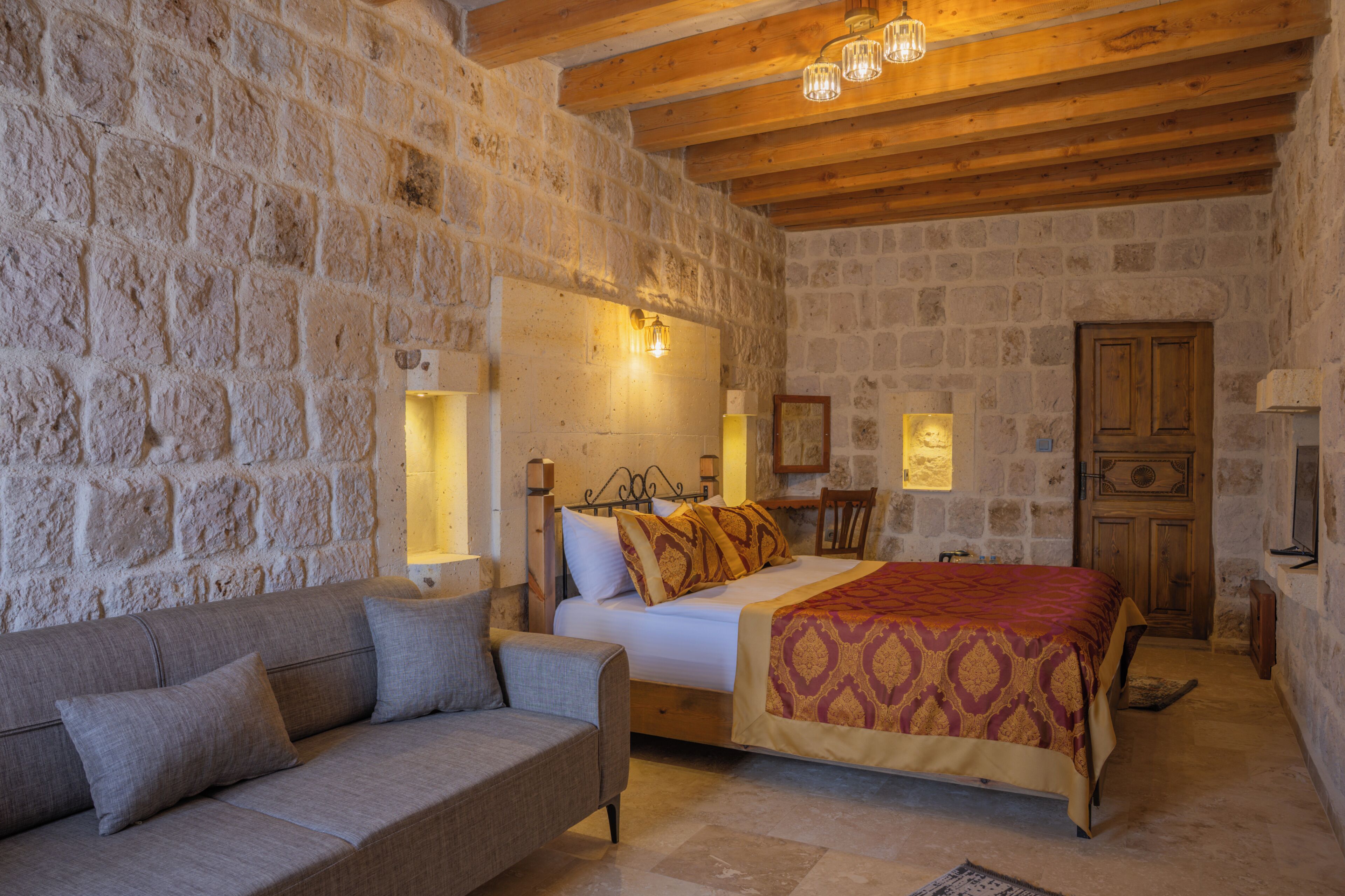 Una habitación con pared de piedra, techo de madera, cama con ropa de cama estampada, sofá y puerta de madera.