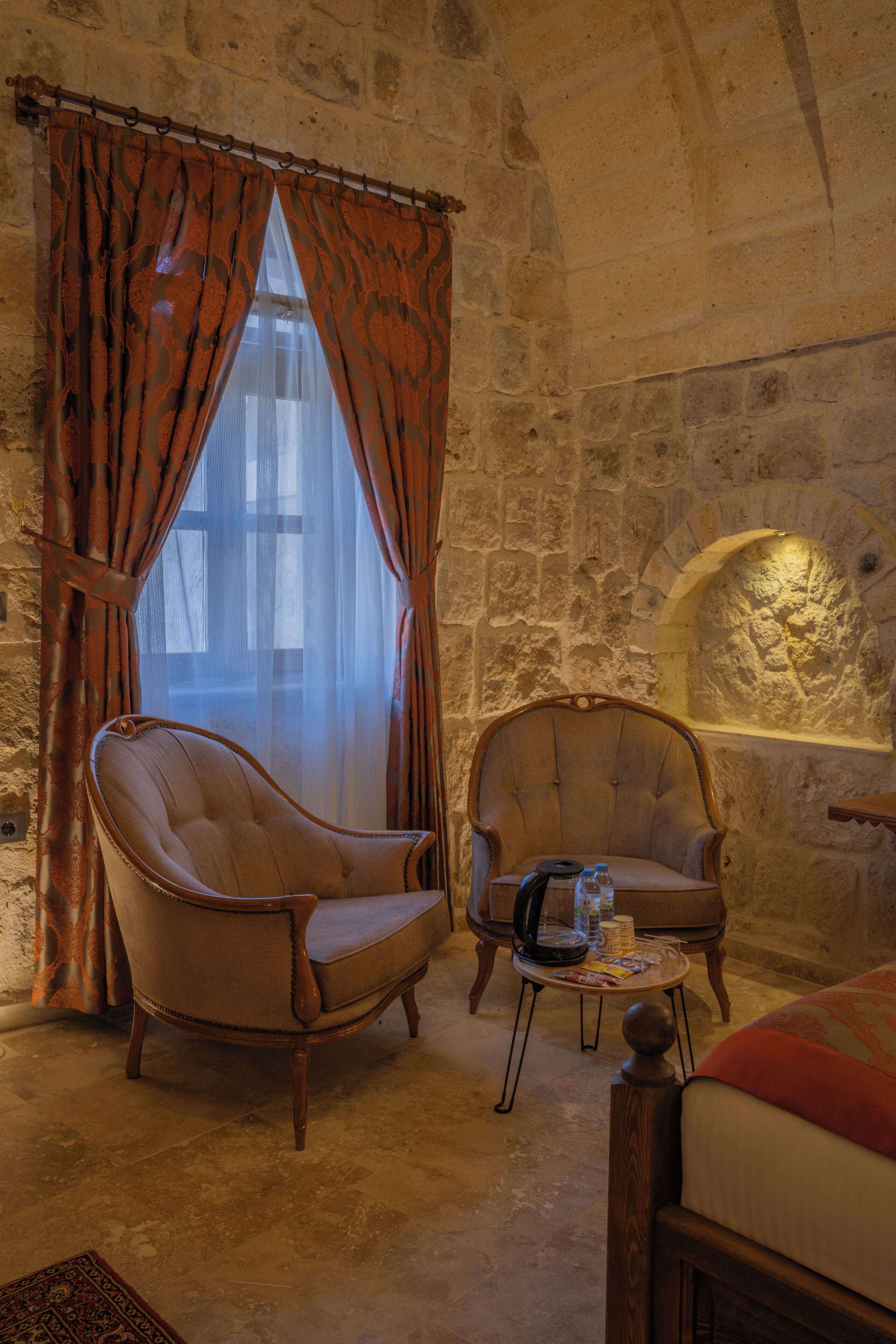 Una habitación con dos sillones, una mesita con objetos, una cama y una pared de piedra con chimenea.