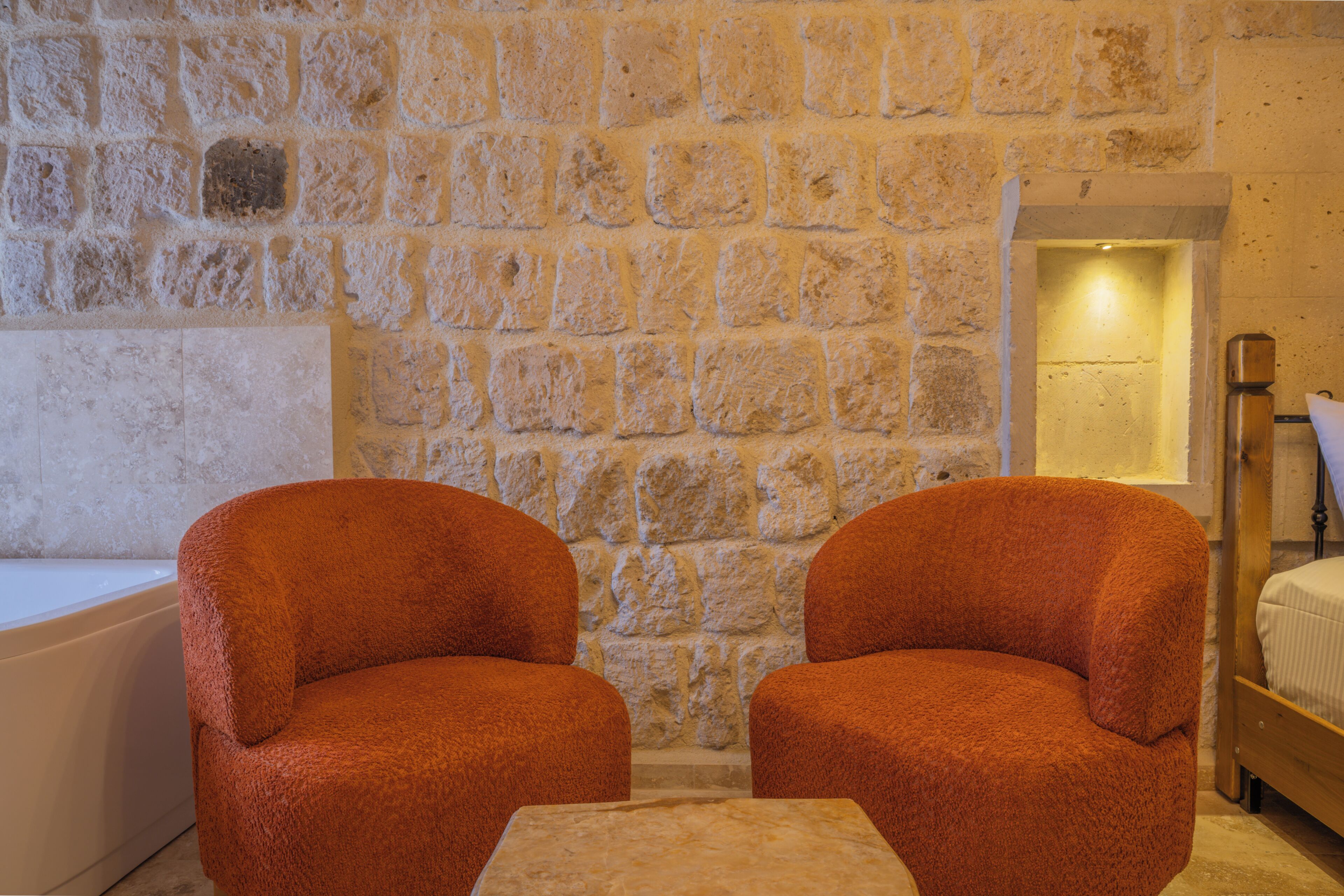 A room with a stone wall, two upholstered chairs, a small table, a bed, and a bathtub.