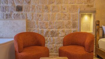 A room with a stone wall, two upholstered chairs, a small table, a bed, and a bathtub.