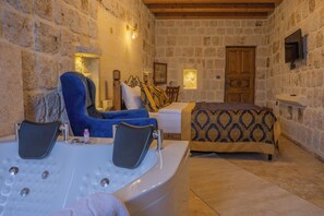 A room with a stone wall, a bed with patterned bedding, a blue armchair, a small table with a bottle, and a bathtub. - Cappa Hisar Cave House (Ortahisar)