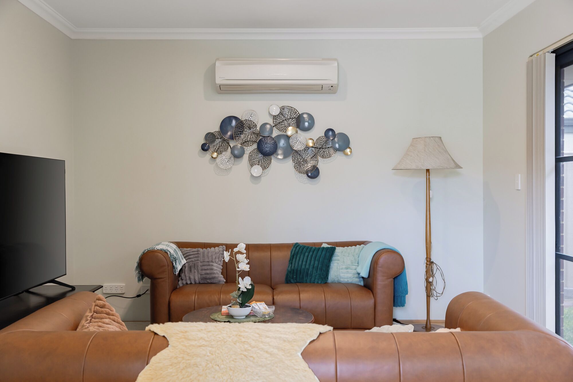 A living room with a brown leather sofa, a flat-screen TV, a wall-mounted air conditioner, decorative wall art, and a floor lamp.