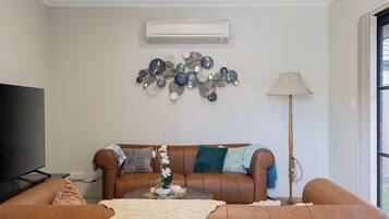 A living room with a brown leather sofa, a flat-screen TV, a wall-mounted air conditioner, decorative wall art, and a floor lamp.