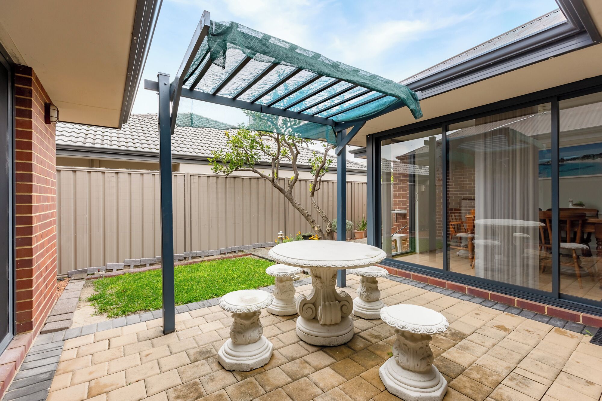 Une terrasse avec une table et quatre tabourets décoratifs, une pergola dotée d’un toit en treillis, un mur en briques et une porte vitrée donnant sur un espace intérieur.