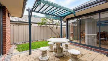 Une terrasse avec une table et quatre tabourets décoratifs, une pergola dotée d’un toit en treillis, un mur en briques et une porte vitrée donnant sur un espace intérieur.