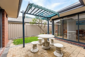 Une terrasse avec une table et quatre tabourets décoratifs, une pergola dotée d’un toit en treillis, un mur en briques et une porte vitrée donnant sur un espace intérieur.