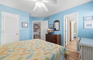 A bedroom with a bed, dresser, and a ceiling fan. There is a door leading to another room and a small table with a chair. - 4/2 bottom floor 1 block from beach (North Myrtle Beach)