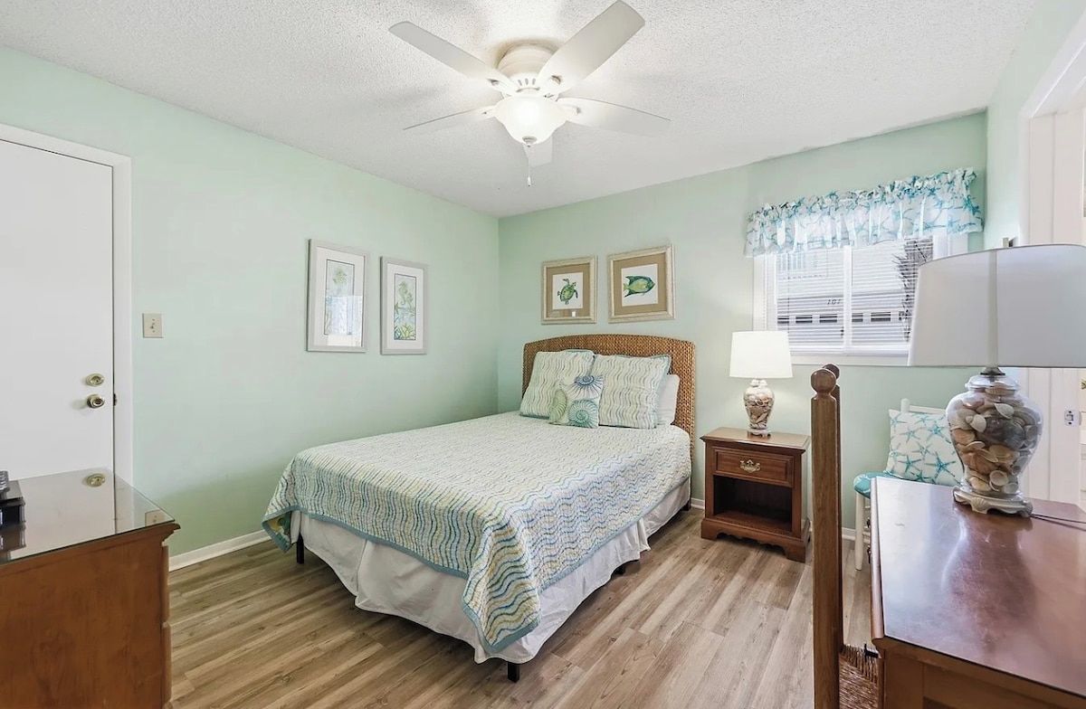 A bedroom with a bed, a nightstand, a lamp, a vase, and a window with blinds.