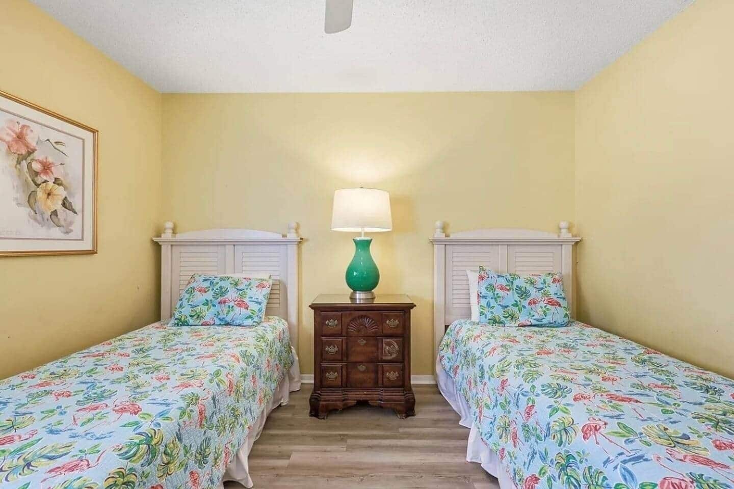 Una habitación con dos camas individuales, una mesita de noche con lámpara y una obra de arte floral enmarcada en la pared.