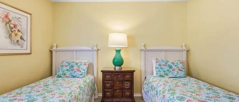 Una habitación con dos camas individuales, una mesita de noche con lámpara y una obra de arte floral enmarcada en la pared.