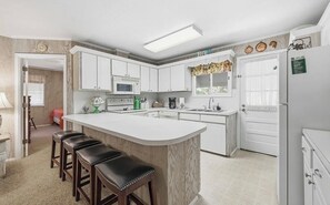Een keuken met witte kasten, een centraal eiland met krukken, een magnetron, een koelkast en een raam met gordijnen.