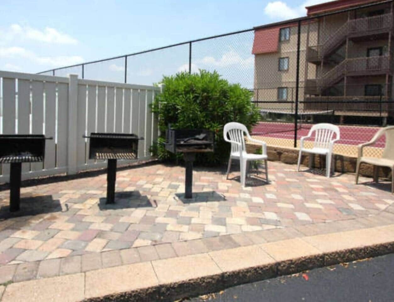 An outdoor patio area with a patterned stone floor, a barbecue grill, a white plastic chair, and a bench.