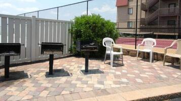 An outdoor patio area with a patterned stone floor, a barbecue grill, a white plastic chair, and a bench.