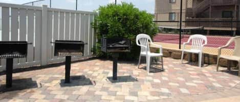 Un espace extérieur aménagé en terrasse, avec un sol en pierre à motifs, un barbecue, une chaise en plastique blanche et un banc.