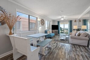 A living room with a blue sofa, white table, and benches, a television, and a view of the outdoors. - Beachfront Escape with Sunset and Marsh Views 14F (North Myrtle Beach)