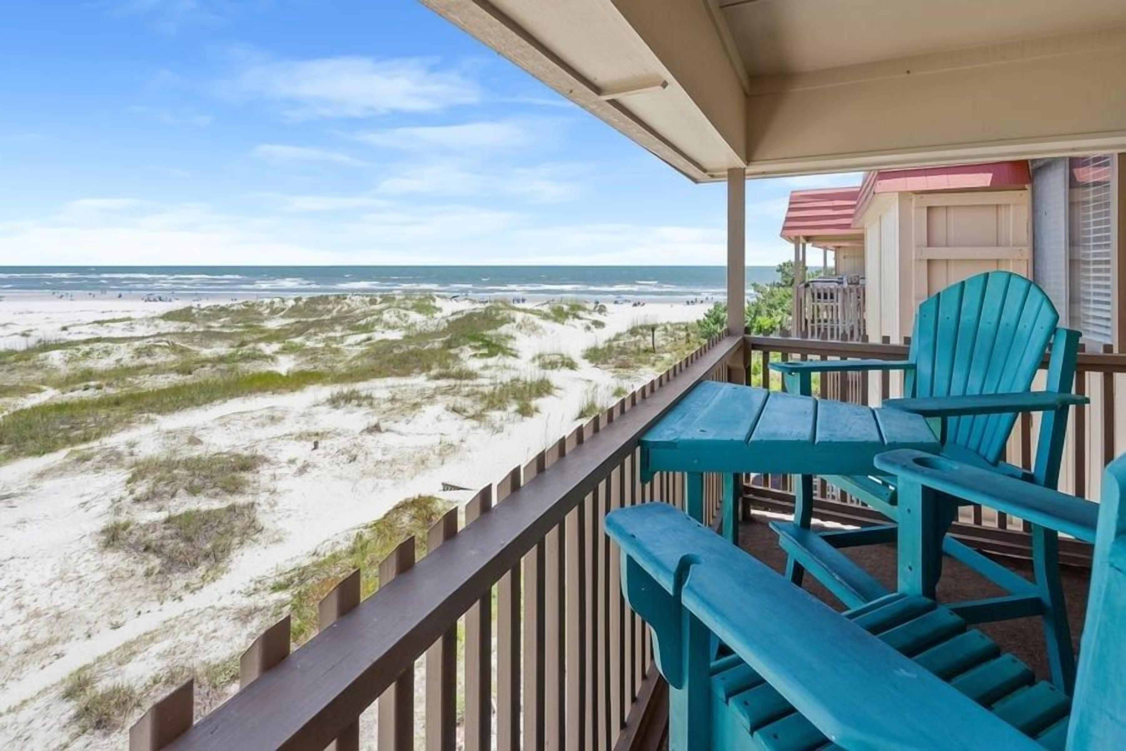 A balcony with a view of the beach, featuring a wooden railing and two blue chairs.