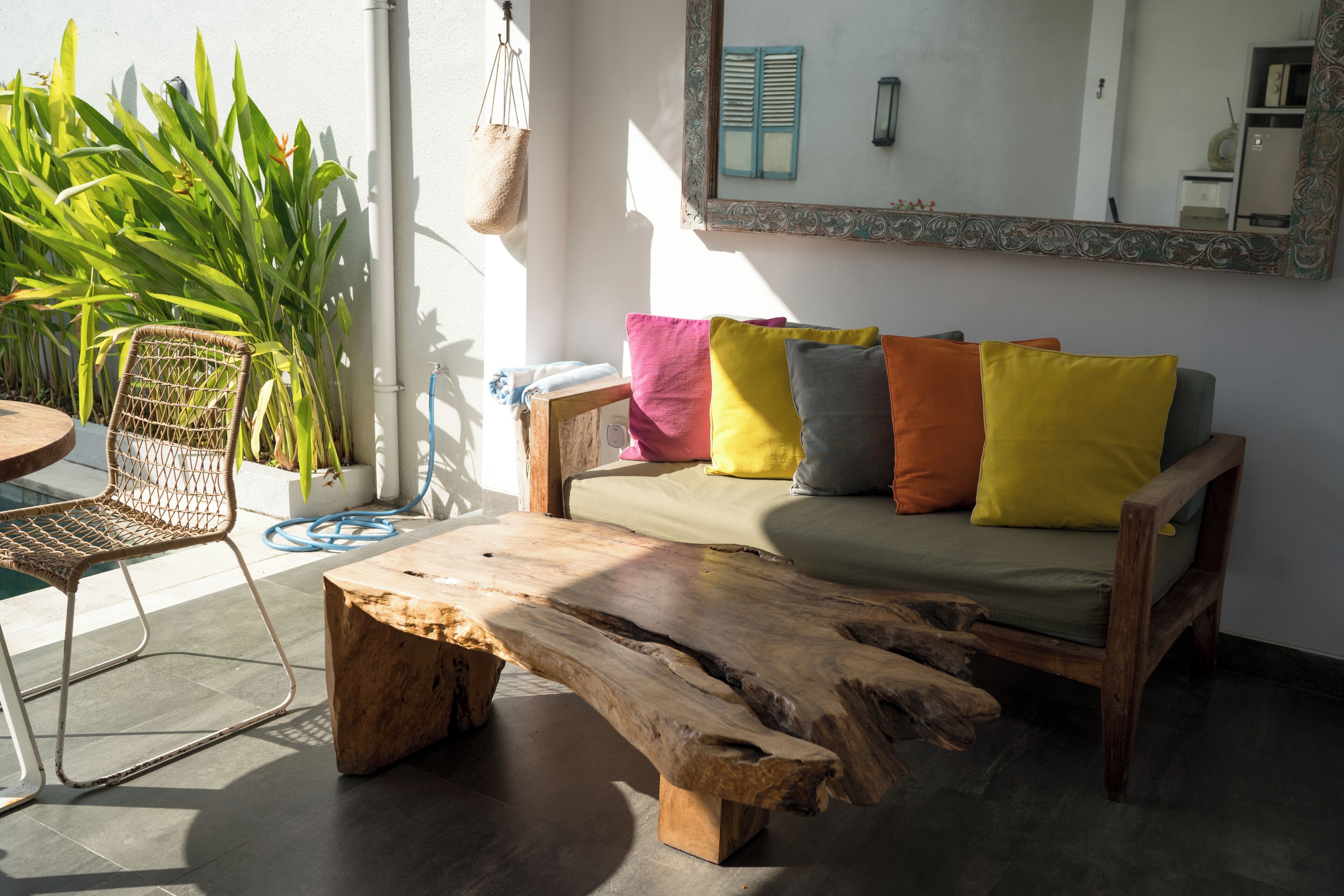 A wooden coffee table, a wicker chair, and a sofa with colorful cushions in a bright room with a large window.