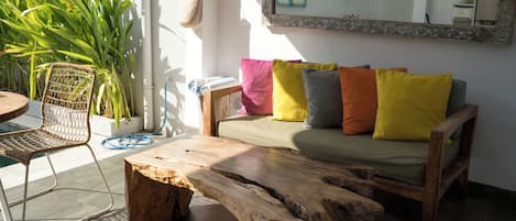 A wooden coffee table, a wicker chair, and a sofa with colorful cushions in a bright room with a large window.