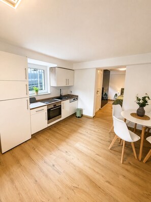 Eine moderne Küche mit weißen Schränken, ein Essbereich mit rundem Tisch und Stühlen sowie ein Blick in ein Schlafzimmer durch eine offene Tür.