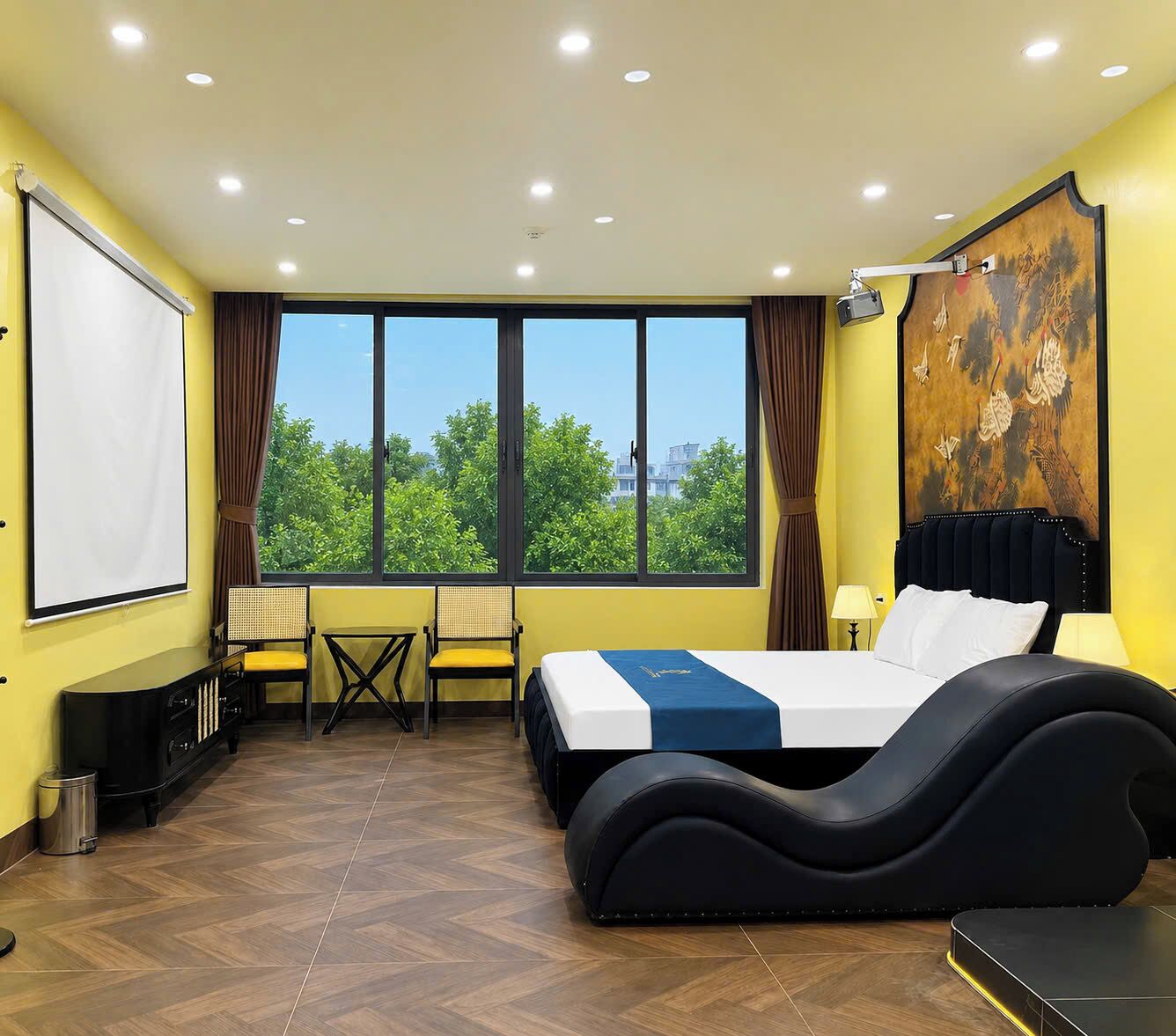 Una camera d'albergo con un letto, una scrivania, una sedia, uno schermo per proiettore e un'ampia finestra vista sul verde.