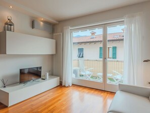 A modern living room with a flat-screen TV, a white sofa, and a balcony with outdoor furniture. - Cosy apartment for 4 people with A/C, WIFI, TV, patio and panoramic view (Riva Del Garda)