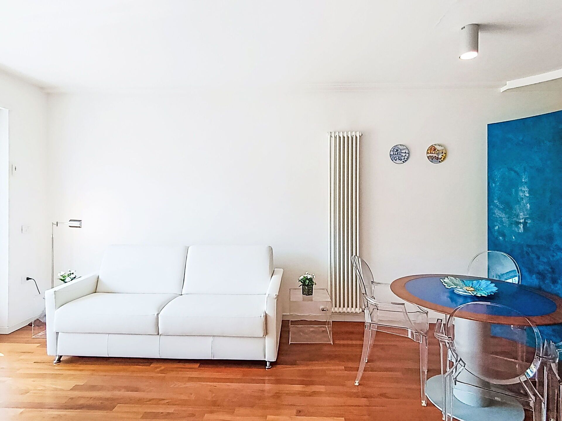 Uma sala de estar moderna com um sofá branco, uma mesa de jantar com cadeiras, um radiador e um quadro abstrato azul na parede.