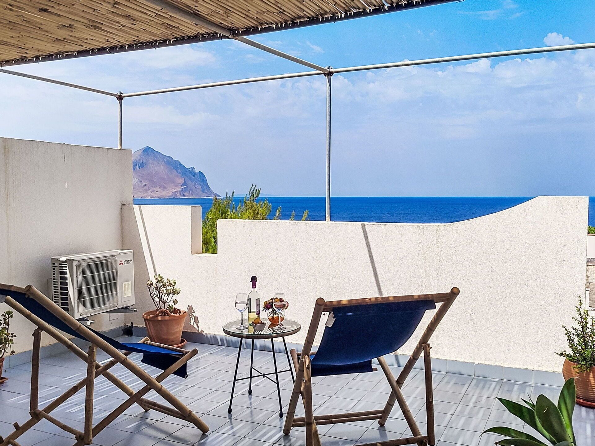 Eine Terrasse mit Blick auf das Meer und einen Berg, mit zwei Stühlen, einem Tisch mit Getränken und einer Topfpflanze.