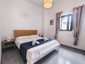 Ein Hotelzimmer mit Bett, Nachttisch, einem fensterbelaubten Fenster und einer Wandlampe.
