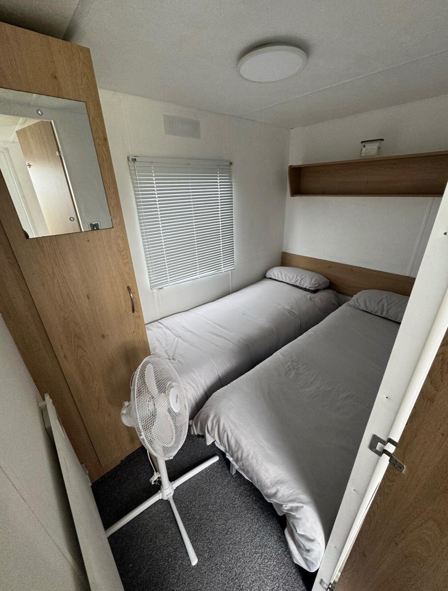 Una caravana pequeña y compacta, de una sola habitación, con cama, un ventilador pequeño y una ventana con persianas.