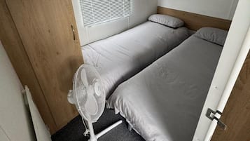 Una caravana pequeña y compacta, de una sola habitación, con cama, un ventilador pequeño y una ventana con persianas.