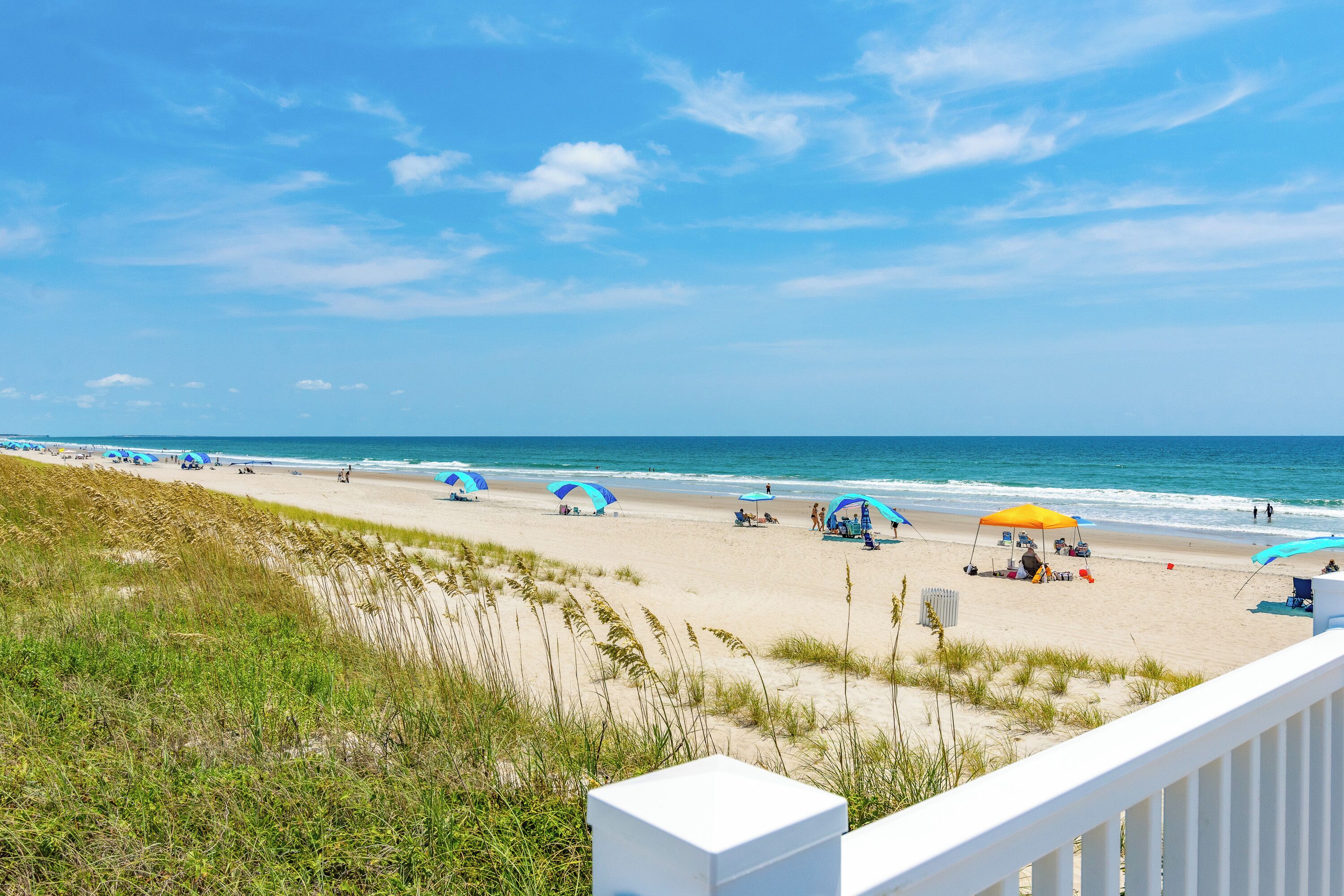 A beach with umbrellas and beach chairs, a grassy area, and a white railing.