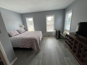 Un dormitorio con cama, cómoda, televisor y dos ventanas.