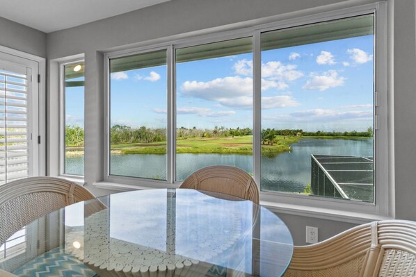 Un comedor con una mesa de vidrio y sillas de mimbre, ventanas amplias con vista a un lago y follaje.