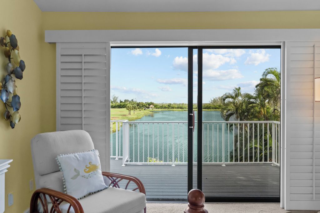 A room with a view of a lake, a chair with a cushion, and a white wall with a decorative wall hanging.