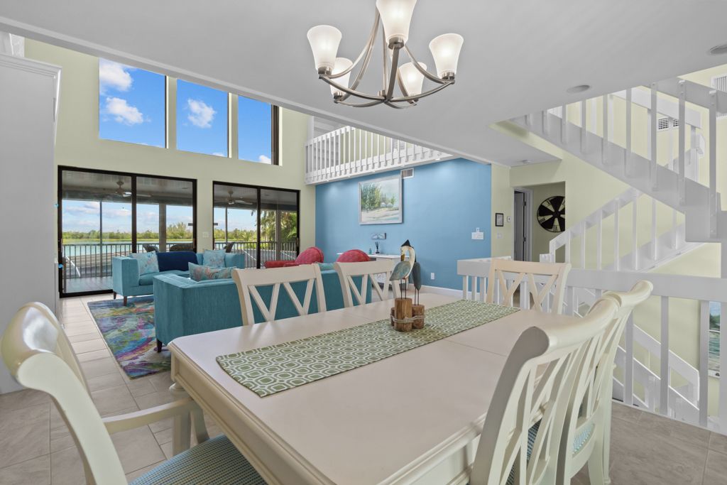 A spacious living area with a dining table, chairs, and a chandelier. There is a staircase with a railing leading to an upper level.